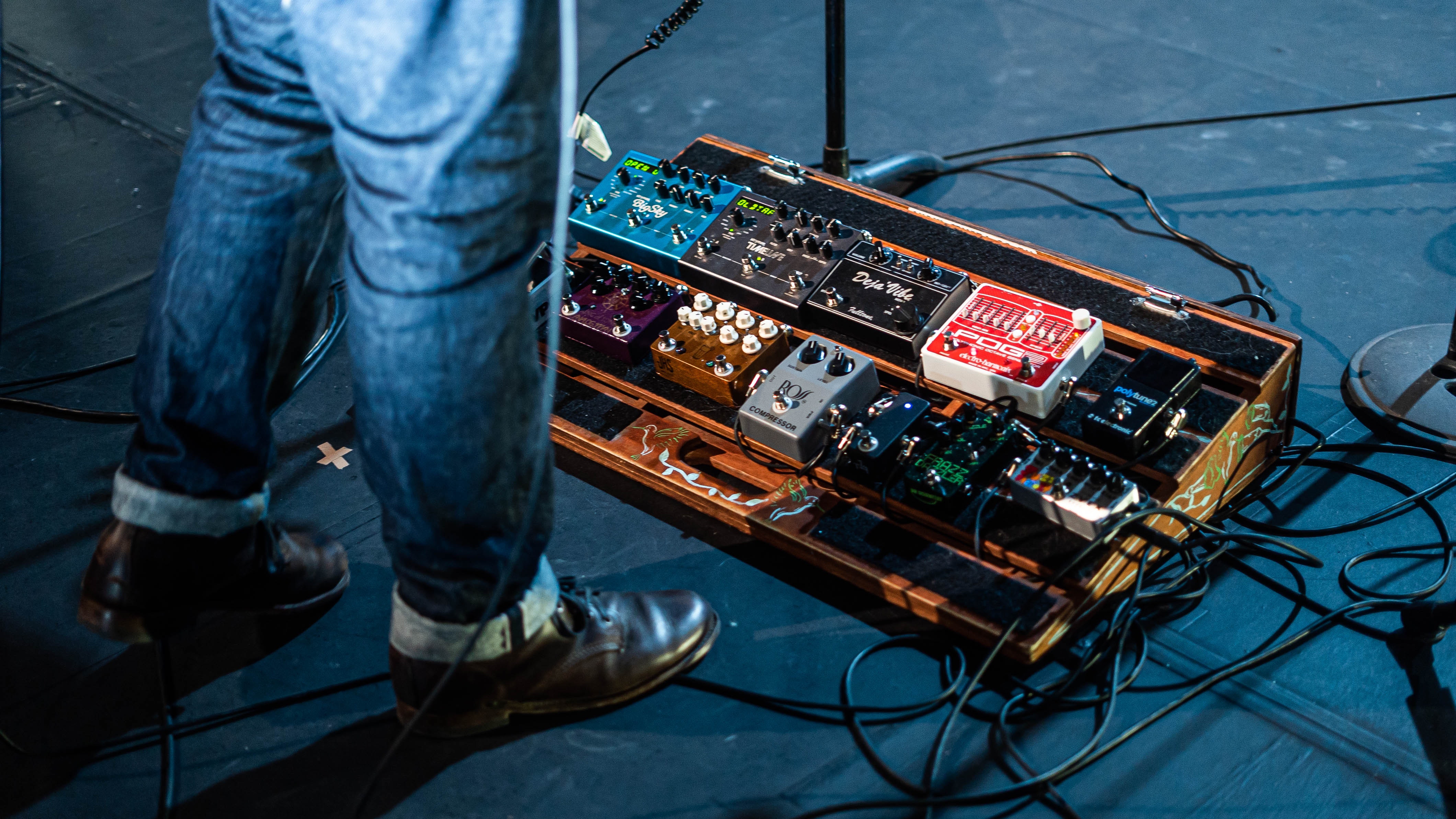 A pedalboard at a man's feet showing a wide variety of guitar effects pedals.