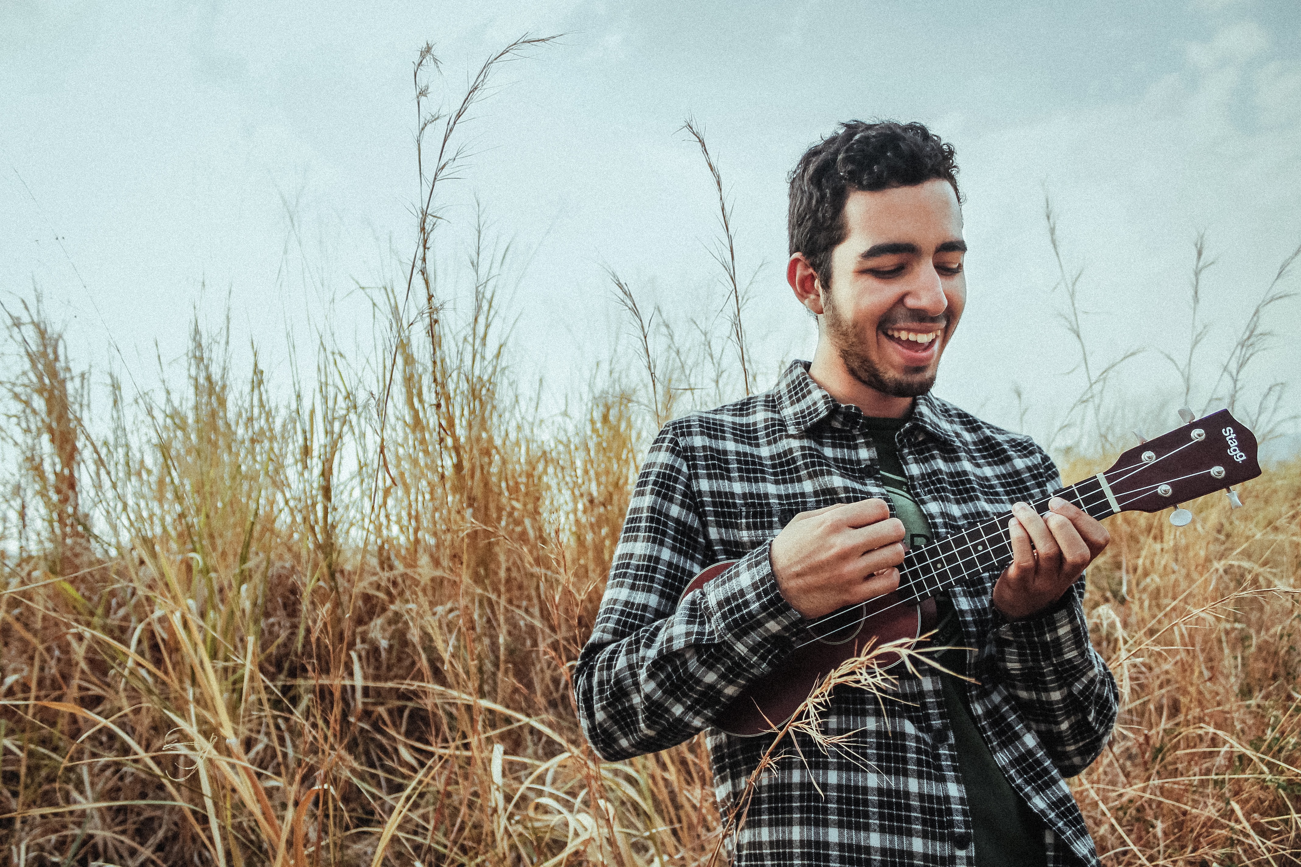 A smiling man playing a ukulele. in a grass field. There are many surprising benefits of playing instruments.